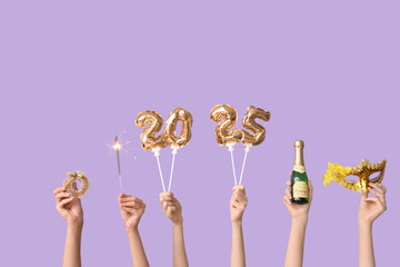 Female hands with figure 2025 made of foil balloons, sparkler, bottle of champagne, Christmas tree decor and carnival mask on lilac background