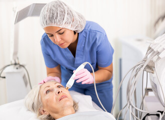 Careful young woman doctor conducting radio frequency lifting procedure on aged woman patient's face