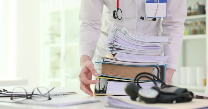 Female doctor lifts heavy stack of ring binders from desk carrying papers away in clinic. Nurse manages paperwork in busy medical environment