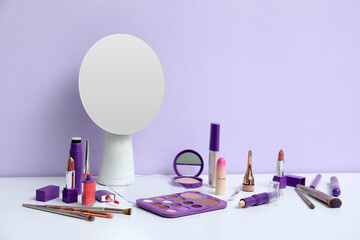 Different decorative cosmetics, makeup brushes and mirror on table near lilac background