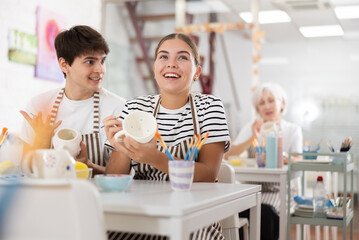 Fototapeta premium Young couple joyfully talking and working together on ceramic pottery, enjoying creative process while painting mugs in light workshop..