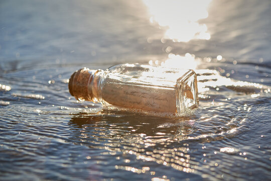 Handwritten letter message in glass on bottle washed up on sand beach with waves and ocean water in the background.