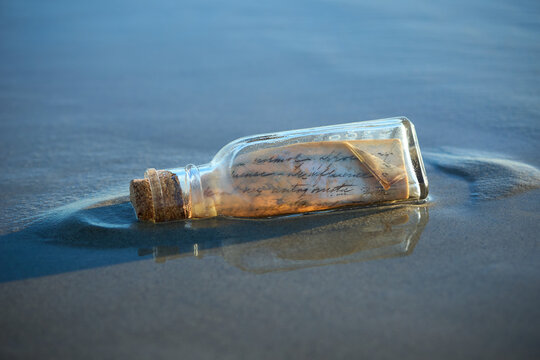 Handwritten letter message in glass on bottle washed up on sand beach with waves and ocean water in the background. 