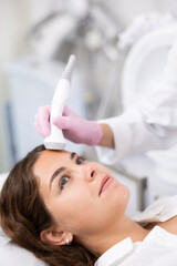 Naklejka premium Faceless cosmetologist doing facial skin ultrasound cavitation for flawless woman face laying in beauty salon indoors