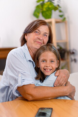 Fototapeta premium Contented grandmother and granddaughter share warm embrace at home, their faces filled with joy and love as they look at camera, capturing heartfelt moment of connection across generations 