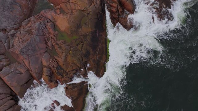 Aerial view of ocean waves crashing against a rocky coastline in Aland, Finland. The red granite cliffs contrast with the dark blue water