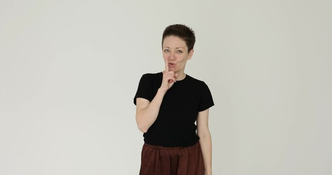 Positive woman with short haircut shows silence gesture on white background. Cunning lady in black t-shirt asks for quiet placing finger on lips