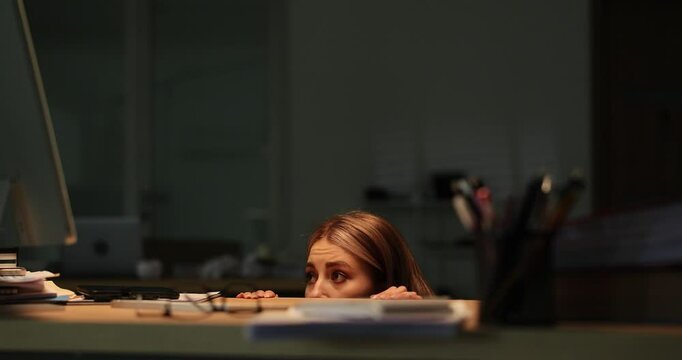 Scared woman hides under desk glancing around nervously in dark office. Employee feels vulnerability and loneliness frightened by thunderstorm
