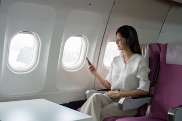 Fototapeta premium Business Woman Traveling in Business Class on a Plane, Using Smartphone and Wearing Headphones, Comfortable and Modern Air Travel