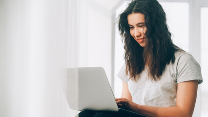 Online shopping. Inspired woman. Digital connection. Smiling satisfied female scrolling laptop sitting near window in light cozy room interior.