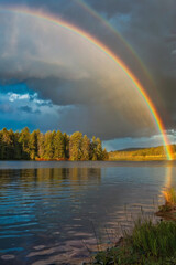 Naklejka premium A vibrant rainbow arches over a serene lake surrounded by misty mountains and lush greenery at dawn