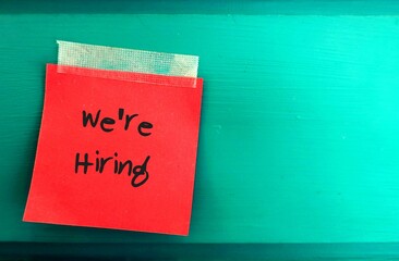 Red paper note with  text written WE'RE HIRING sticking on the green board wall with blank copy...
