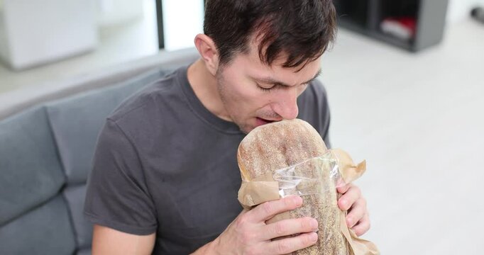 Man enjoys aroma of fresh bread sitting on sofa in office room. Hungry guy eats pastry enjoying delightful taste taking moment to rest slow motion
