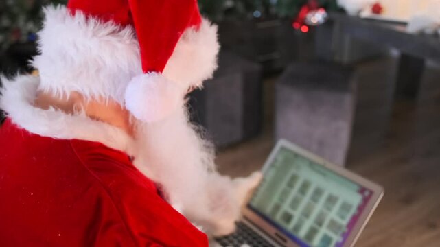 Close up of charming Santa Claus looking at camera and laughing ho-ho-ho using laptop browsing folders and pages on internet, back side view.