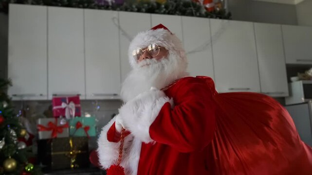 A white-bearded Santa in glasses and with a bag of gifts on his shoulders comes out of a room decorated with a Christmas tree, festive decorations and New Year's garlands.