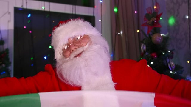 Santa Claus with gray beard and glasses hold Italian flag, dances next to Christmas tree and twinkling garlands. Slow motion.
