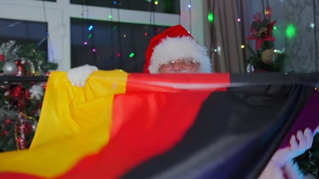 Stylish Santa dancing with flag of Germany and heart-shaped gift box against background of colorful garlands and sparkling Christmas tree.