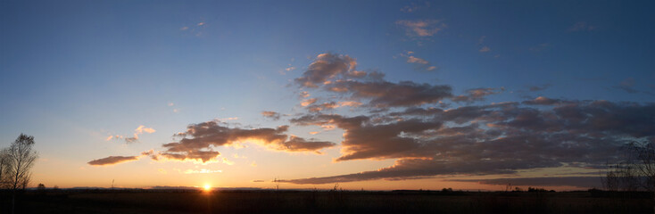 Fototapeta premium Sun sets low on horizon, casting golden glow across landscape. Fluffy clouds, illuminated with warm hues, float against clear blue sky. Serene scene tranquil, with last light of day fading gently.