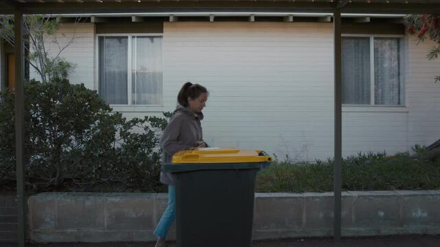 A woman in a gray jacket pulls a yellow-lid garbage bin along a pathway outside a white suburban house