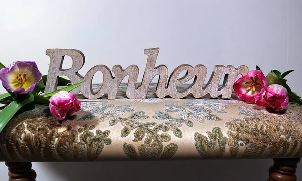 Sculpture en bois du mot fran&ccedil;ais "Bonheur" sur un coussin brod&eacute;, d&eacute;cor&eacute;e de fleurs roses, symbolisant la joie et l'&eacute;l&eacute;gance
