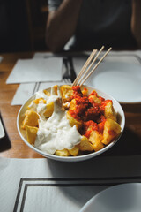 White bowl full of fried potatoes covered in aioli and spicy tomato sauce sits on a wooden restaurant table
