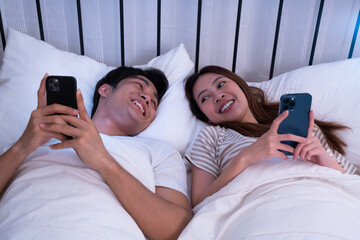 Asian young couple lying in bed, smiling and holding smartphones for browsing the internet, enjoying each other company in a relaxed and cozy at night home