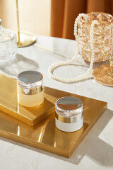 Two skincare mockups are arranged on two copper platforms, surrounded by a necklace, glass cups, and other decorations. The luxurious backdrop image template is perfect for promoting beauty products.
