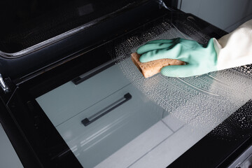 Woman in green gloves cleaning oven door with sponge in modern kitchen, closeup.