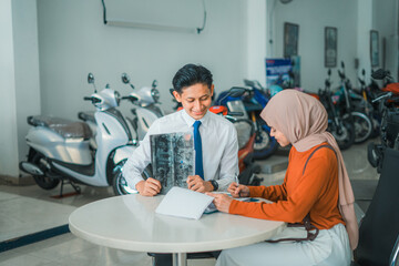 motorcycle sales man serves a veiled female customer while sitting looking at brochures and price lists in a showroom