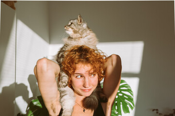 portrait red-haired curly young woman with pet gray fluffy cat in arms, lit by bright sun