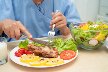 Asian senior woman patient eating pork chop stake and vegetable salad for healthy food in hospital.