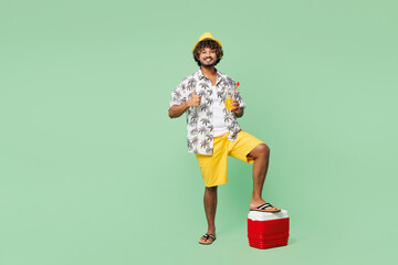 Full body young Indian man he wear beach shirt hat rest near hotel pool put leg on cooler fridge drink orange cocktail walk go isolated on plain green background. Summer vacation sea sun tan concept.