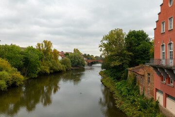 Fototapeta premium river runs through a town with a bridge over it. The water is calm and the trees lining the riverbank are lush and green. The scene has a peaceful and serene atmosphere