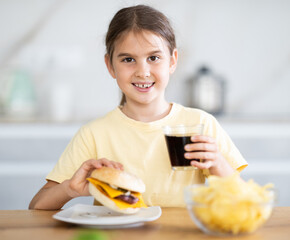 Little girl eating meat burger and drinking carbonated drink in kitchen