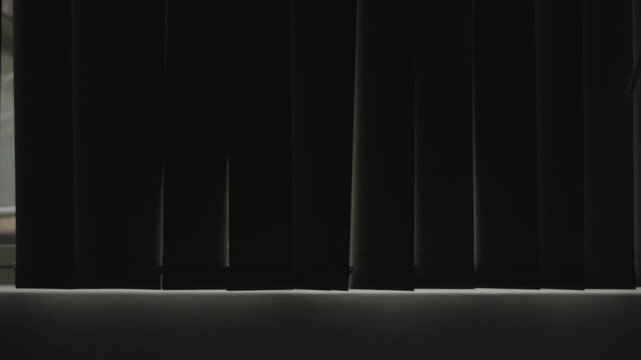 Dark room with blackout shades moving slowly showing green trees in background. Window rolling or sliding modern vertical blinds in closed house or apartment. Daylight revealed outside behind fabric