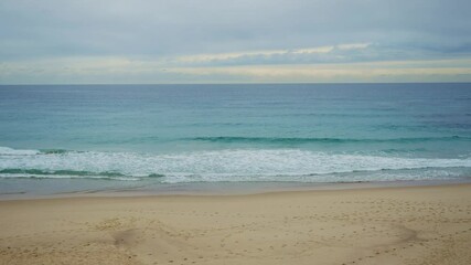 Wide shot empty beach with footprints on sand and small waves in ocean in winter. Cold temperature and winds outdoors in a Sydney beach during wintertime. No people in quiet and cloudy afternoon