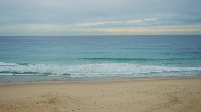 Wide shot empty beach with footprints on sand and small waves in ocean in winter. Cold temperature and winds outdoors in a Sydney beach during wintertime. No people in quiet and cloudy afternoon
