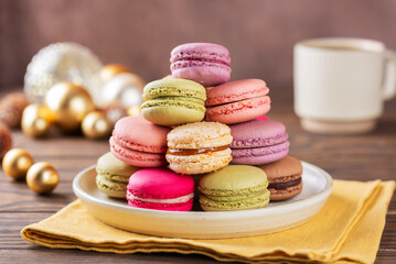 Multicolored macaroons in a plate with cup of coffee and Christmas balls on a yellow napkin. Christmas background.