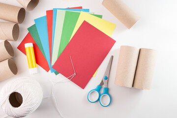 Craft materials for paper decorations, featuring colored paper sheets, cardboard tubes, glue stick, scissors, needle, and white thread. Flat lay composition for DIY and crafting projects