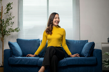 Young asian woman sitting on couch smiling looking away from camera
