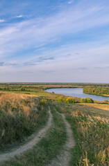 A road winds through a grassy field with a river in the background