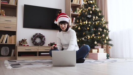 Young Caucasian woman wearing Santa hat and headphones enjoying music near Christmas tree. Festive atmosphere with decorations and gifts creating joyful holiday mood. - Powered by Adobe