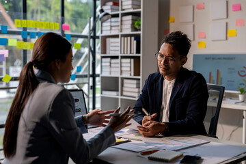 A woman and a man are talking in a room with a lot of papers on the table