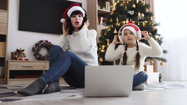 Caucasian young adult mother and little daughter sing songs while listening to carols in headphones using a laptop. Family leisure during the festive season against the background of Christmas tree.