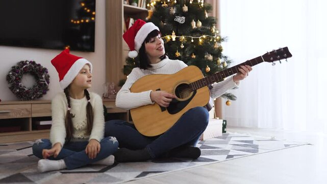 Asian mother and daughter wearing Santa hats enjoy festive holiday time singing Christmas carols while playing guitar. They sit together near decorated Christmas tree with lights and ornaments.