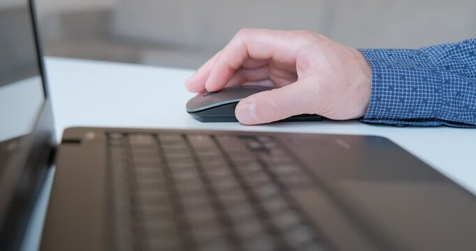 An individual is engaged in work, using a computer mouse to navigate a laptop in a contemporary workspace. The setting is bright and organized, reflecting productivity.