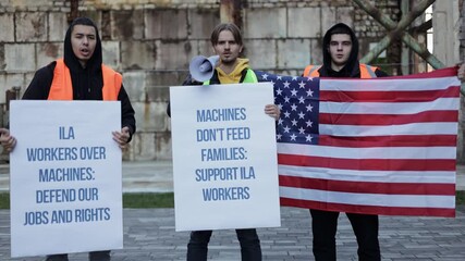 Dockworkers protest against automation and job losses, holding signs and the American flag. They advocate for workers' rights and job security. - Powered by Adobe