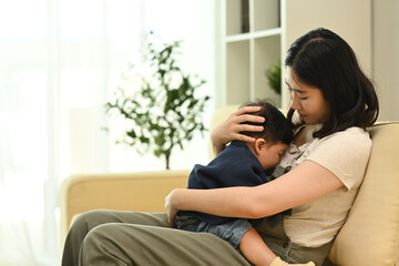 Young mother sitting on sofa, gently cradling her baby in her arms. Parenting, child care and family concept