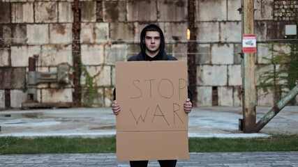 A young man holds a sign reading Stop War in front of a dilapidated building, conveying a message of peace and protest against conflict.