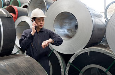 A man in a hardhat and mechanic coverall dust protection suit, talks on his phone while checking time on his watch. He stands near large rolls of metal in metal sheet manufactory.